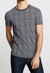 Krátký rukáv tričko v námořnické látce s květinovým vzorem v bílé barvě. Má kulatý výstřih s kontrastním lemem.