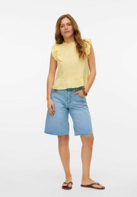 Top giallo in broderie anglaise con maniche voluminose, abbinato a pantaloni corti in denim azzurro e infradito marroni. Design semplice e minimalista.