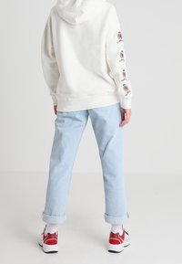 Felpa bianca con cappuccio e ricami sulle maniche, jeans azzurri a vita bassa e scarpe da ginnastica con dettagli rossi, che mostrano un look casual.