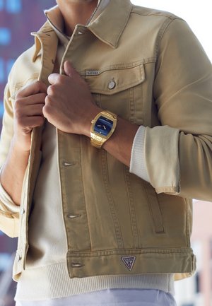 Homme portant une veste beige Guess et un pull, tenant le col de la veste avec les deux mains, montre dorée avec cadran bleu au poignet gauche.