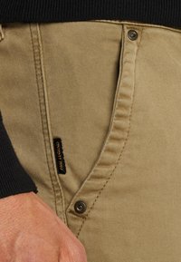 Pantalons kaki avec une texture lisse, dotés d'une ceinture noire, d'un bouton en métal et d'une petite étiquette noire avec "Paul Legend" sur le côté.