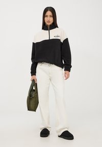 Pullover in pile nero e crema con mezza zip, polsini ed elastico in vita. Abbinato a jeans bianchi a gamba larga e una borsa verde.