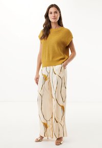 Sweter w kolorze musztardowym z teksturowanym wzorem, zestawiony z zwiewnymi kremowymi spodniami z złotymi i czarnymi abstrakcyjnymi liniami oraz złotymi sandałami na odkrytej podeszwie.