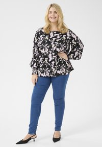 Svart och rosa abstrakt mönstrad blus med ballongsärmar, ihop med blå tajta jeans och svarta slingback klackar, stående mot en ensartad bakgrund.