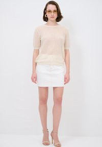 Beige gestricktes Oberteil mit kurzen Ärmeln und einem strukturierten Muster, kombiniert mit einem weißen Jeans-Minirock. Die Accessoires umfassen gelb getönte Sonnenbrillen und Sandalen.