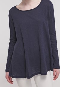 Granatowa bluzka z długimi rękawami o szerokim dekolcie, o luźnym kroju i lekkim, teksturowanym materiale. Posiada zaokrąglony dół i boczne rozcięcia.