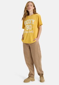 Gele oversized t-shirt met witte grafische tekst, gecombineerd met lichtbruine linnenbroek. Schoenen zijn beige veterschoenen.