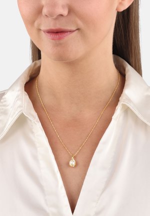 Collar de cadena de oro con un colgante irregular y brillante. Llevado sobre una blusa de satén blanca, resaltando su superficie reflectante y diseño delicado.