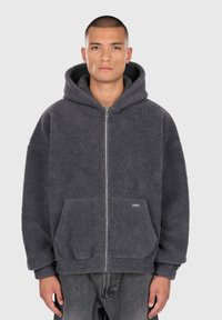 Mand med kort hår iført en mørkegrå lynlås-hoodie af fleece med frontlommer og løse ærmer, stående foran en ensfarvet baggrund.