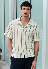 Short-sleeve, striped shirt in cream with dark green lines, featuring a button-down front and a relaxed collar, made of textured fabric.