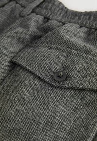 Grauer, strukturierten Stoff mit einer sichtbaren Tasche und einem schwarzen Knopfdetail. Der Stoff hat ein gewebtes Hahnentrittmuster, ist weich und mittelschwer.