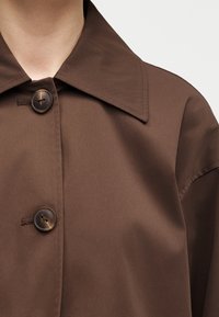 Καφέ παλτό με γιακά και δύο μεγάλα καφέ κουμπιά στο μπροστινό μέρος, φορεμένο από ένα άτομο, εμφανίζοντας το πάνω μέρος του στήθους και της περιοχής των ώμων.