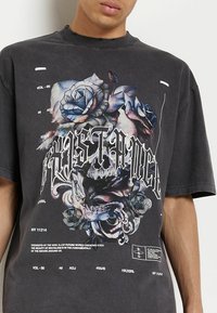 Camiseta de gran tamaño en gris oscuro con un gráfico central de rosas, un cráneo y un texto en negrita. El material parece suave con un ajuste holgado y cuello redondo.