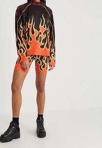 Černý top s dlouhým rukávem a oranžové šortky s plamenným vzorem; elastická tkanina; doplněné o robustní černé platformové boty.