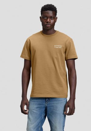 Mann trägt braunes T-Shirt mit dem Text "WORKWEAR IS OUR SOUL" auf der linken Brust und blaue Jeans, steht vor einem schlichten hellen Hintergrund.