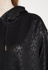 Sudadera negra de tejido texturizado con un sutil patrón floral. Detalle de cordón con un terminal de metal en el cuello.