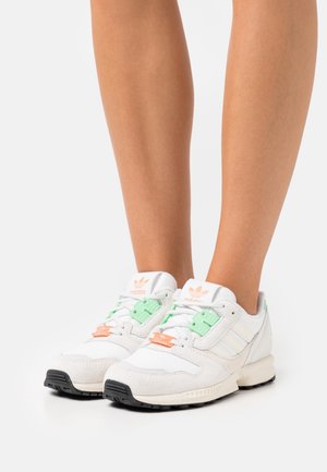 Baskets blanches Adidas avec des accents verts pastel et orange portées sur des jambes nues sur un fond blanc uni.