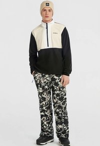 Melna un krēmkrāsas krāsu bloku puszipa virsjaka ar logotipu; apvienota ar brīvi pieguļošām melnām biksēm ar baltu traipu rakstu.