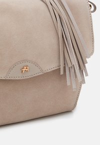 Beżowa zamszowa torebka na ramię z klapką, ozdobiona jasnymi frędzlami oraz małym, złotym, wytłoczonym logo na przedniej stronie. Gładka faktura.