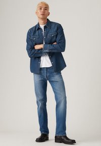 Donkerblauwe denim shirt met westernstijl yoke, twee voorzakken en drukknoopsluiting, gecombineerd met lichtblauwe jeans en zwarte boots.