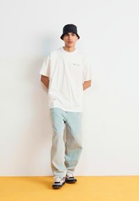 Hvid t-shirt med et lille print, lyseblå jeans og sort bucket hat. Sneakers med sort, hvidt og gråt accent. Står op ad en blank væg.