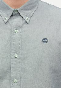 Ανοιχτό γκρι πουκάμισο με κουμπιά, γιακά και μικρό σκούρο μπλε λογότυπο δέντρου κεντημένο στην αριστερή πλευρά του στήθους.