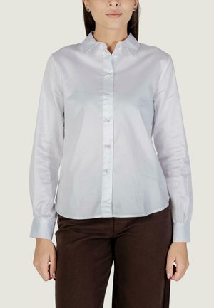 Donna che indossa una camicia bianca a maniche lunghe con bottoni e pantaloni marroni, in piedi contro uno sfondo chiaro con le mani rilassate lungo i fianchi.
