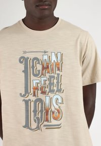Camiseta de algodón beige de manga corta, con un texto gráfico multicolor que dice "PUEDO SENTIR A LOIS" y un diseño de flecha encima del texto.