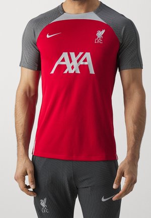 Mężczyzna w czerwono-szarej koszulce piłkarskiej Liverpoolu i szarych szortach z białymi logo Nike i LFC.