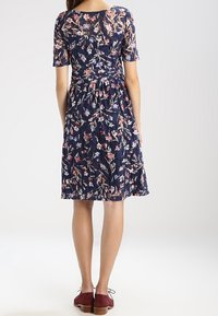 Marineblaues Blumenkleid mit kurzen Ärmeln, aus strukturiertem Stoff und tailliert, verziert mit rosa und weißen Blumenmustern. Braune Schuhe.