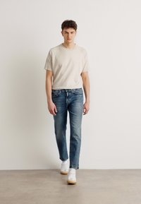Tricou din bumbac bej cu mâneci scurte, asortat cu blugi albaștri slim-fit. Modelul poartă adidași albi. Design simplu și modern.