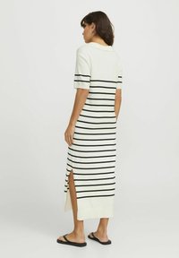 Vestido a la altura de la rodilla, de manga corta, en color crema con rayas horizontales negras. Presenta aperturas laterales y un cuello clásico para un detalle adicional.