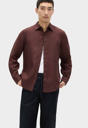 Mann trägt ein braunes Hemd über einem weißen T-Shirt und schwarzen Hosen, steht mit den Händen in den Taschen vor einem schlichten Hintergrund.