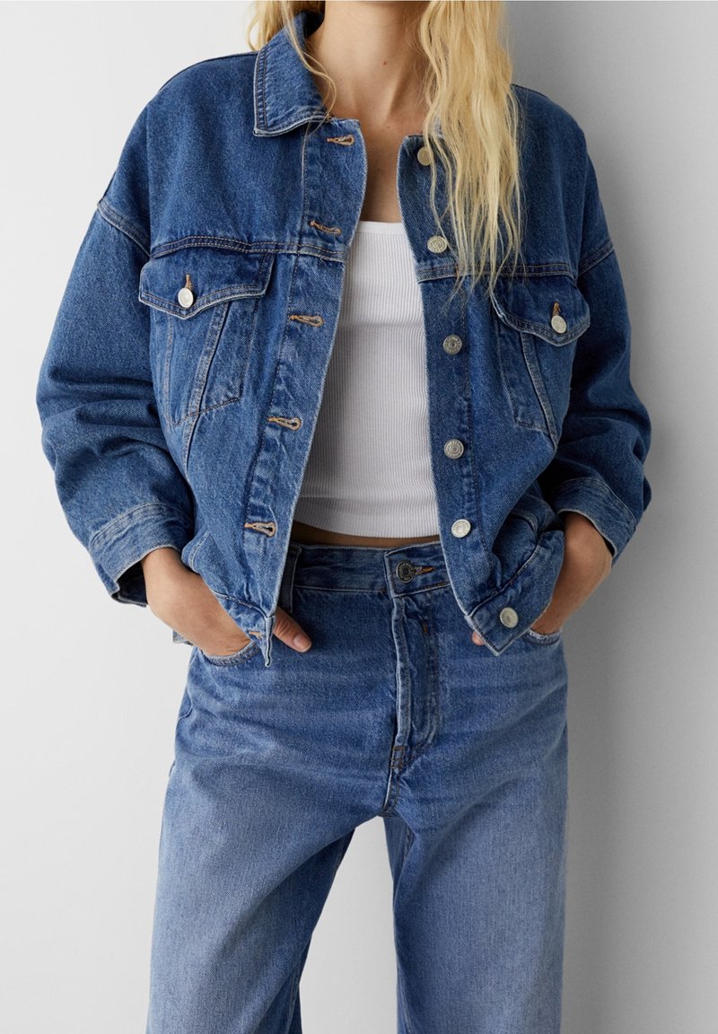 Personne portant une veste en jean bleu et un jean avec les mains dans les poches, associé à un top court blanc côtelé.
