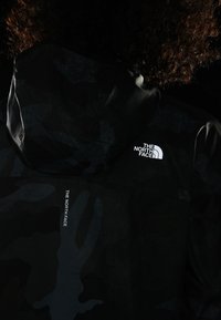 Chaqueta de camuflaje negra con capucha, que presenta un logo blanco de "The North Face" en el hombro y una textura suave y resistente al agua.