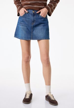 Kobieta w niebieskiej mini z denimu, brązowej bluzce w paski z długim rękawem, białych skarpetkach oraz ciemnobrązowych trampkach z beżowymi sznurowadłami.