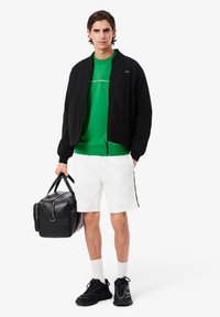 Sort bomberjakke over en grøn sweatshirt, hvid shorts og sorte sneakers. Holder en sort duffeltaske med sølvhardware.