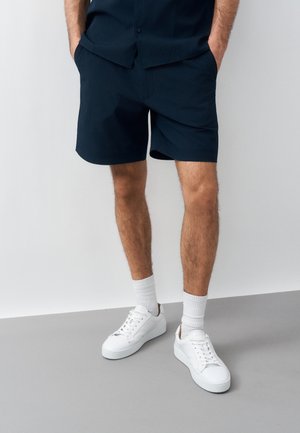 Homme portant un short bleu marine, des chaussettes blanches montantes et des baskets blanches, debout les mains dans les poches devant un fond uni.