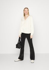Blusa longa de mangas com decote em V na cor marfim, combinada com calças pretas de corte flare. Os acessórios incluem uma bolsa texturizada preta e sapatos brancos de slip-on.