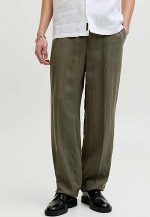 Une personne portant un pantalon olive ample, des chaussures habillées noires et une chemise blanche, avec une main dans une poche et des bracelets à un poignet.