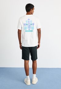 White cotton t-shirt with colorful graphic print on the back, featuring blue and purple designs. Paired with dark shorts and sneakers.