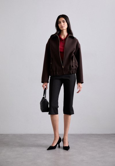 Braune Zip-Jacke mit hohem Kragen, kombiniert mit einer roten Bluse mit Knöpfen, schwarzen cropped Hosen und schwarzen High Heels. Hält eine schwarze Handtasche.