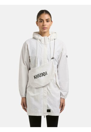 Frau trägt eine weiße Kapuzenjacke und eine weiße Umhängetasche mit dem Logo "KHUJO 2001" und steht vor einem schlichten hellen Hintergrund.
