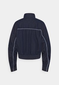 Mörkblå denimjacka med hög krage, elastisk midja och vit piping längs baksidan och ärmarna, sedd bakifrån.