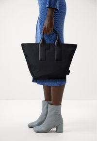 Donna in un vestito blu a trama che tiene una grande borsa nera, indossando stivali alla caviglia grigi chiari con tacco a blocco su uno sfondo bianco.