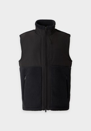 Homme portant un gilet noir sans manches sur une chemise blanche à manches longues, pantalon noir et baskets blanches, debout devant un fond uni.