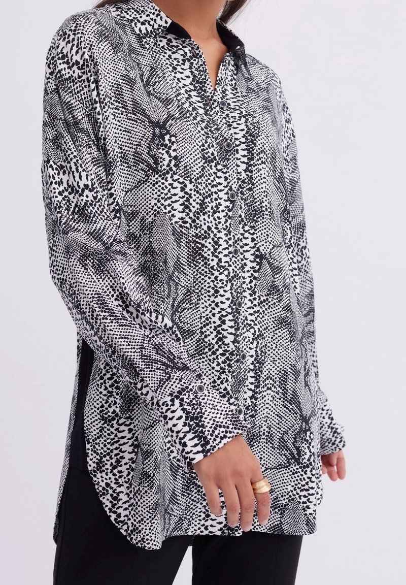 Femme portant une chemise à manches longues boutonnée avec un imprimé animal abstrait noir et blanc, associée à un pantalon noir et des bagues dorées à la main gauche.