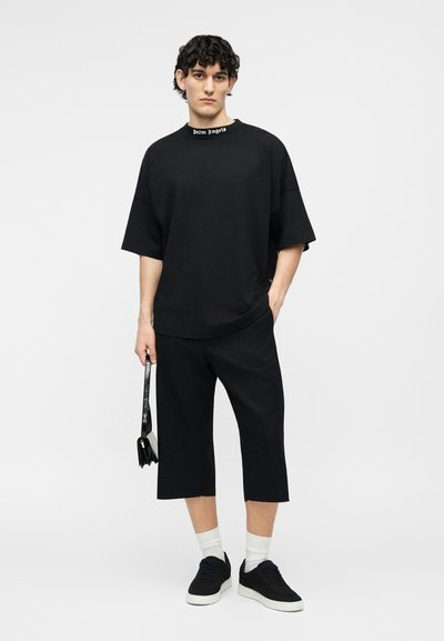 Tânăr purtând o cămașă neagră supradimensionată cu textul „Palm Angels” pe guler, pantaloni negri scurtați, adidași, ținând o geantă mică neagră.