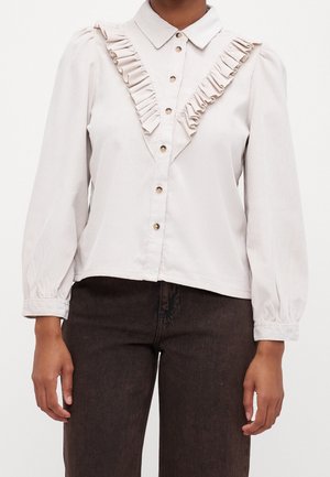 Kobieta w kremowej bluzce zapinanej na guziki z falbanką z przodu i długimi rękawami, w połączeniu z ciemnymi spodniami.