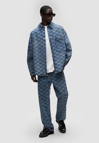 Completo di giacca e pantaloni in denim blu chiaro con motivo ripetuto del logo, dal fit rilassato, con bottoni e tasche laterali. Indossato con una t-shirt bianca.